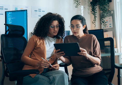 Two women sit in desk chairs looking at a tablet device. 